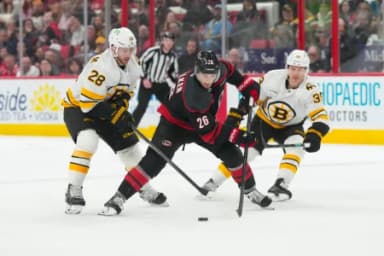 Carolina Hurricanes defenseman Sean Walker (26) skates with the puck between Boston Bruins center Elias Lindholm (28) and center Morgan Geekie (39) during the third period at Lenovo Center.